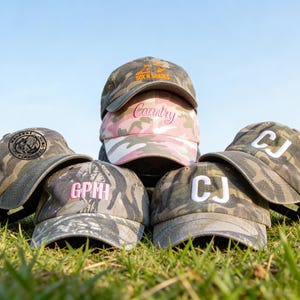 May include: A collection of six baseball caps featuring various camouflage patterns. The caps display embroidered text including "SOX N SHADES," "Country," "GPM," and "CJ" in different colours and fonts, with one cap showing a circular fishing logo.