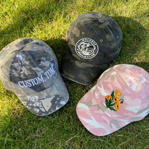 May include: Three baseball caps with camouflage patterns. One grey cap features the white embroidered text CUSTOM TOM. One dark grey cap displays a circular white patch with the text MALAKAI FISHING CLUB. One pink cap features an embroidered floral design.