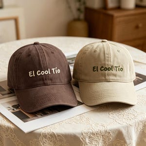 Op de afbeelding: Twee baseballpetten, één bruine en één beige, liggen op een tafel. Elke pet heeft een gebogen klep en de geborduurde tekst "El Cool Tío" in het wit. De petten zijn gemaakt van een zachte stof.