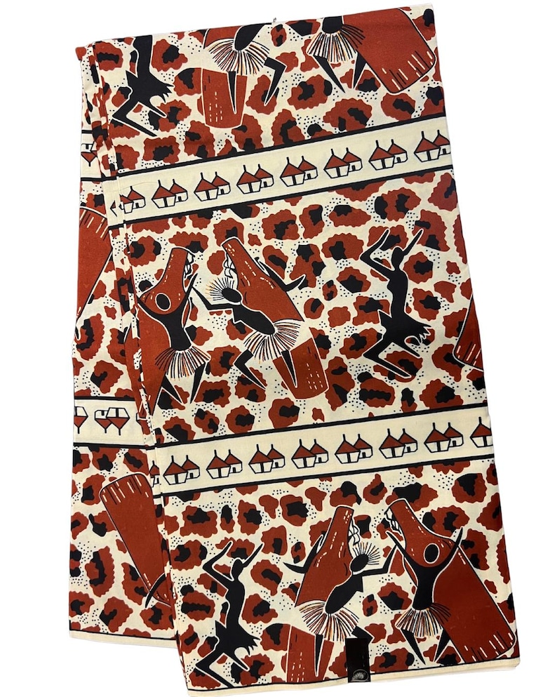 Op de afbeelding: Een witte stof met een bruin en zwart patroon van dansende figuren en huizen. De stof heeft een luipaardprint achtergrond.