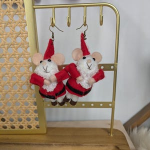 Op de afbeelding: Een paar rode en witte vilt muis oorbellen met kerstmanmutsen. De oorbellen hangen aan een gouden oorbellenstandaard.