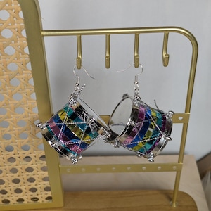 Puede incluir: Un par de pendientes de plata con forma de pequeños tambores. Los tambores están decorados con cuadrados multicolores iridiscentes y tienen detalles plateados.
