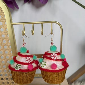 Op de afbeelding: Een paar cupcake-vormige oorhangers met witte frosting, rode en witte snoepstokken, roze en groene pompoenen en een bruine fluwelen cupcake-basis.