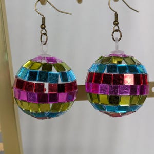 Może przedstawiać: Para kolczyków w kształcie kuli dyskotekowej. Każdy kolczyk ma kulę pokrytą małymi, kolorowymi, odblaskowymi kwadratami w odcieniach niebieskiego, zielonego, różowego i czerwonego. Kolczyki wiszą na złotych haczykach.