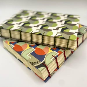 Puede incluir: Dos diarios hechos a mano con cubiertas de patrones geométricos. El diario superior tiene un patrón verde, marrón y blanco. El diario inferior tiene un patrón rojo, naranja, amarillo, azul y verde.
