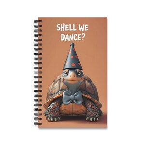 Op de afbeelding: Een spiraalgebonden notitieboek met een bruine omslag waarop een cartoon schildpad te zien is met een zwart-rode stippenpartyhoed en een blauwe strik. De tekst "Shell We Dance?" is in wit op de omslag gedrukt.