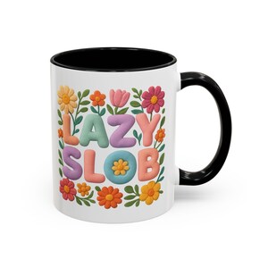 May include: White ceramic mug with a black interior and handle. The mug features the words "LAZY SLOB" in colorful, 3D-style letters, surrounded by a floral design with orange, pink, yellow, and green accents.