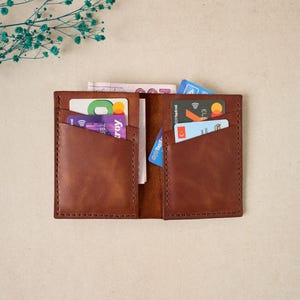 May include: Open brown leather wallet with multiple card slots and a central compartment. The wallet is filled with various credit cards and paper currency. The stitching is visible around the edges, and the wallet is displayed on a neutral background.