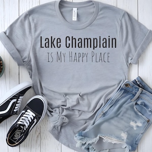 May include: Gray t-shirt with the text "Lake Champlain is my happy place" printed on the front. The shirt is laid flat on a white wooden surface with a pair of black and white sneakers and a pair of light blue denim shorts.