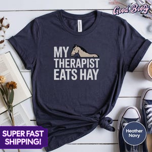 Op de afbeelding: Een marineblauw t-shirt met een witte afbeelding van een paard en de tekst "My Therapist Eats Hay".