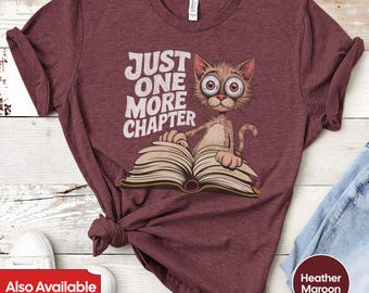May include: A maroon t-shirt with a cartoon cat wearing glasses and reading a book. The text on the shirt says "Just One More Chapter".