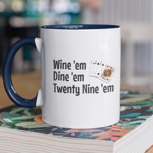 May include: White ceramic coffee mug with a blue rim and handle. The mug has a black graphic that says "Wine 'em Dine 'em Twenty Nine 'em" with a picture of playing cards.