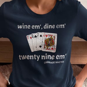 May include: A navy blue t-shirt with the text "wine em', dine em'" and "twenty nine em'" printed on it. The shirt also features a graphic of five playing cards, including a Jack of Hearts, a 5 of Clubs, a 5 of Spades, and two 5 of Diamonds. The text "CRIBBAGE MASTER" is printed below the graphic.