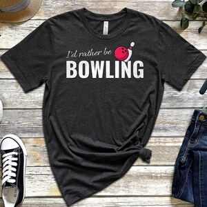Peut inclure: Un t-shirt gris foncé avec le texte "I'd rather be bowling" et un graphique de quille de bowling blanche et de boule de bowling rouge.