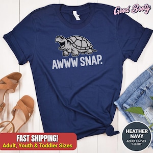 May include: Navy blue unisex t-shirt featuring a cartoon turtle with the text "AWWW SNAP." The shirt is made of heather navy material and is available in adult, youth, and toddler sizes. The shirt has rolled-up sleeves.