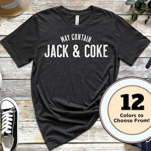 May include: Dark gray t-shirt with the text "MAY CONTAIN JACK & COKE" in white. The image also shows black sneakers, a straw hat, glasses, a watch, and jeans. A circular graphic indicates 12 colors to choose from.