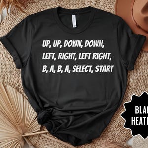 May include: Black heather t-shirt with white text: "UP, UP, DOWN, DOWN, LEFT, RIGHT, LEFT RIGHT, B, A, B, A, SELECT, START." The shirt is displayed on a woven surface with a hat and decorative elements.
