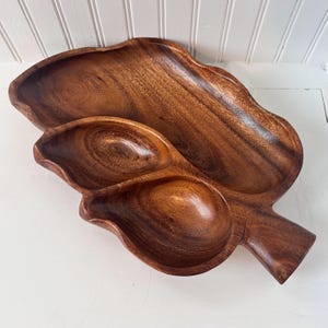 May include: A wooden serving tray with a leaf design. The tray is made of a dark brown wood and features three separate compartments. The largest compartment is oval-shaped, and the other two are smaller and round. The tray has a handle.