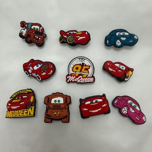 May include: A set of ten colourful rubber shoe charms featuring various characters and symbols from the Cars movie franchise, including Lightning McQueen, Mater, and Sally, along with a circular emblem displaying the number 95 and text.