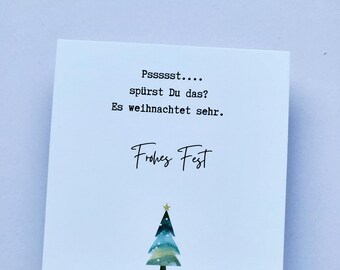 May include: A white square card with black text and a watercolor Christmas tree illustration. The text reads "Psssst... spürst Du das? Es weihnachtet sehr. Frohes Fest."