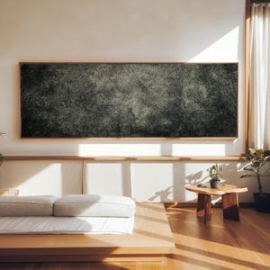 Puede incluir: Una gran obra de arte abstracta rectangular con una superficie oscura y texturizada está enmarcada en madera clara. La obra de arte cuelga sobre una cama baja con ropa de cama blanca. También se ven una pequeña mesa auxiliar de madera y plantas en macetas.
