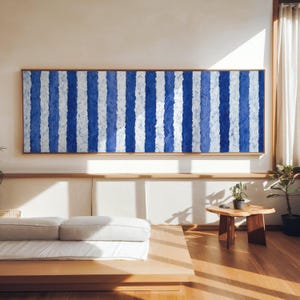 Peut inclure: Une grande peinture abstraite rectangulaire avec des rayures verticales bleues et blanches dans un cadre en bois. L'œuvre est exposée au-dessus d'une plate-forme en bois basse avec un matelas et des oreillers blancs. La pièce a des planchers en bois et une petite table en bois.
