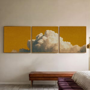 Peut inclure: Trois œuvres d'art murales carrées encadrées. Chaque pièce présente un fond jaune moutarde texturé avec une formation de nuages au centre. Les nuages sont blancs et gris, créant un contraste avec le jaune.