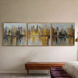 Può includere: Un trittico di dipinti astratti incorniciati raffiguranti un paesaggio urbano con una superficie d'acqua riflettente. L'opera presenta una tavolozza di toni oro, marrone e grigio, creando un effetto strutturato e impressionistico. I dipinti sono esposti sopra una panca marrone.