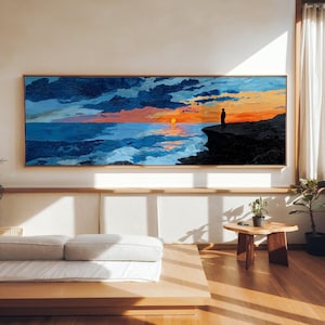 Peut inclure: Une grande peinture rectangulaire représente un coucher de soleil sur l'océan. Le ciel est un mélange de bleus, d'oranges et de jaunes, avec une silhouette sur une falaise. L'œuvre est encadrée de bois clair.