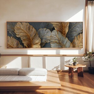 Peut inclure: Grande œuvre d'art rectangulaire représentant des feuilles stylisées dans des tons or et gris sur un fond sombre. L'œuvre est encadrée de bois clair, apportant une touche d'élégance inspirée de la nature à la pièce. Mesure environ 150 cm de long.