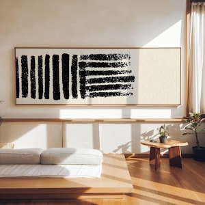 May include: Large rectangular abstract art piece with a light beige background and bold black brushstrokes. The artwork is framed in a thin, natural wood frame and hangs on a light-colored wall. The piece adds a modern touch to the room.