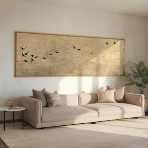Peut inclure: Un canapé beige avec plusieurs coussins est placé sous une grande œuvre d'art rectangulaire. L'œuvre présente un fond doré avec des silhouettes d'oiseaux noirs. Une petite table d'appoint et une plante en pot sont également visibles.