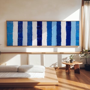 May include: A large, framed abstract painting featuring vertical stripes in varying shades of blue and white. The artwork is displayed in a bright room. The painting's textured surface is evident. The frame is a natural wood colour.
