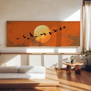 Pode incluir: Uma obra de arte em tela retangular retrata um bando de pássaros pretos em silhueta, voando sobre um grande sol amarelo contra um fundo laranja e marrom. A obra de arte é emoldurada em uma moldura de madeira natural fina.