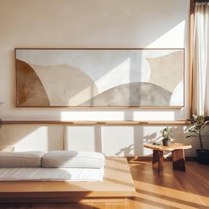 Può includere: Un grande dipinto astratto rettangolare in una cornice di legno. L'opera presenta forme morbide e curve in tonalità beige, marrone e grigio. Il dipinto è appeso sopra una piattaforma bassa in legno con un materasso e cuscini bianchi. Un tavolino di legno e piante in vaso sono nella stanza.