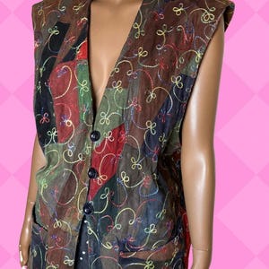 Può includere: Un gilet senza maniche con un design patchwork nei toni del marrone, rosso, verde e nero. Il gilet presenta uno scollo a V, tre bottoni scuri e colorati motivi floreali ricamati. Ha due tasche anteriori.