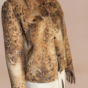 May include: A brown and tan leopard print jacket with a collared neckline, two chest pockets, and button closures. The jacket is worn over a white skirt. The jacket's pattern is a repeating design of dark brown spots.