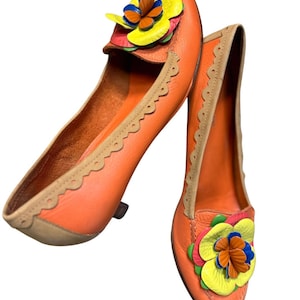 Op de afbeelding: Een paar oranje leren ballerina's met een bloemversiering. De ballerina's hebben een geschulpte rand en een beige binnenkant. De bloem is gemaakt van geel, blauw, rood en oranje leer.