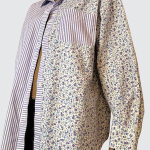 Può includere: Una camicia a maniche lunghe con un design patchwork. La camicia presenta una combinazione di tessuto a righe verticali bianche e lavanda e una stampa floreale con fiori blu e viola su sfondo bianco. Ha un colletto e una tasca.