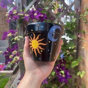 Könnte beinhalten: Ein schwarzer Terrakotta-Pflanztopf mit einer handgemalten gelben Sonne mit geschlossenen Augen und einem hellblauen Halbmond. Das Design ist mit kleinen, bunten Punkten verziert, die Sterne auf der dunklen Oberfläche darstellen.