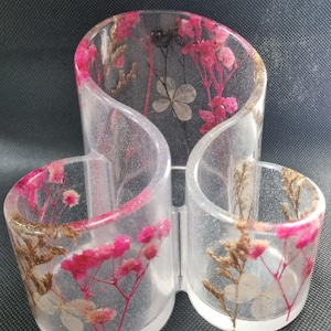 Può includere: Un organizer da scrivania in resina trasparente con tre scomparti collegati. La resina è incorporata con fiori secchi rosa e bianchi e steli color oro. L'organizer ha un aspetto glitterato e scintillante ed è posto su uno sfondo scuro.