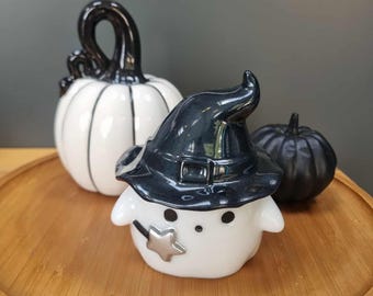 May include: A Halloween display featuring a ceramic ghost in a witch's hat, a white pumpkin with black detailing, and a small black pumpkin. The items are arranged on a wooden tray, creating a festive arrangement.