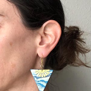 May include: Hand-painted wooden earrings in a triangular shape. The earrings feature a design of a yellow sun over blue and white waves. The earrings are suspended from gold-colored ear wires. The earrings are approximately 2 inches in length.