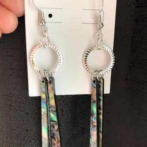 May include: A pair of silver-toned earrings with a textured circular design and iridescent rectangular pendants. The earrings have a hook closure and are displayed on a white card.