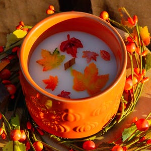 Peut inclure: Une bougie en céramique orange avec un intérieur en cire blanche, décorée de feuilles d'automne colorées et d'un bâton de cannelle. La bougie est posée dans une couronne de baies rouges et de feuilles vertes, créant une esthétique automnale chaleureuse.