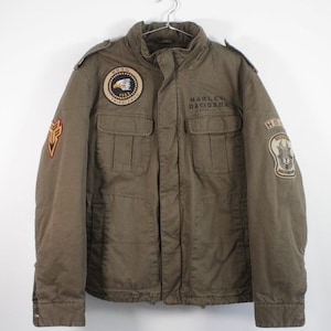Può includere: Giacca Harley-Davidson verde oliva con toppe multiple e logo Harley-Davidson stampato sul petto. La giacca ha una chiusura con bottoni, tasche multiple e un colletto alto. Le toppe presentano aquile e altri emblemi.
