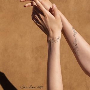 Puede incluir: Dos tatuajes florales simples en los brazos de una persona. Los tatuajes son dibujos de l&iacute;nea negra de una vid con peque&ntilde;as flores. Los tatuajes est&aacute;n en las mu&ntilde;ecas y los antebrazos.