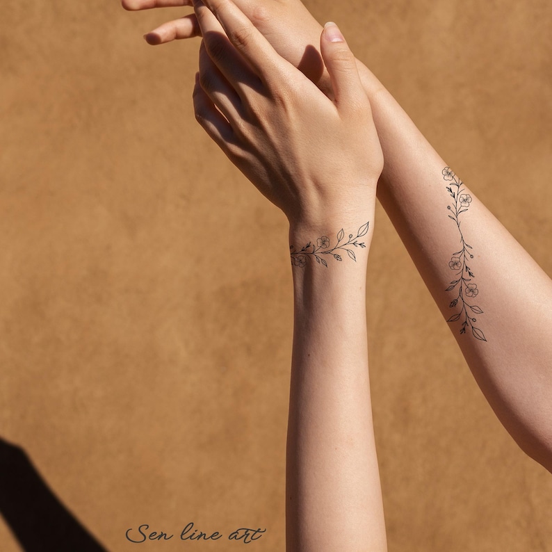Puede incluir: Dos tatuajes florales simples en los brazos de una persona. Los tatuajes son dibujos de l&iacute;nea negra de una vid con peque&ntilde;as flores. Los tatuajes est&aacute;n en las mu&ntilde;ecas y los antebrazos.