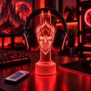 May include: A black gaming headset rests on a clear acrylic stand featuring an illuminated red skull design. The stand has a textured, circular base with a power button and is accompanied by a small remote control with multiple colour buttons.