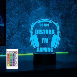 Puede incluir: Una luz nocturna LED azul con forma de auriculares con el texto "No molestar, estoy jugando". La luz est&aacute; sobre una base negra y tiene un mando a distancia.